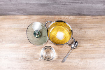 A metal pan with a glass lid contains millet grits, salt, sugar and a mug of water for cooking millet porridge on a wooden table