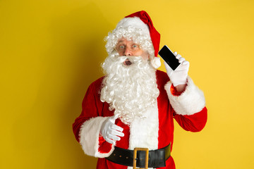Santa Claus with modern gadgets isolated on yellow studio background. Caucasian male model in traditional costume. Concept of Christmas, New Year's, winter mood, technologies. Shows empty phone screen