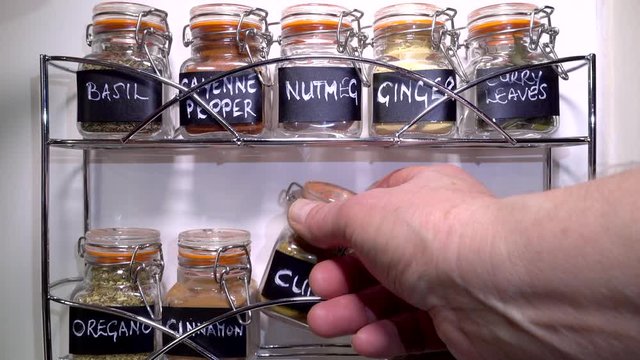 Slow motion close POV shot of a man&rsquo;s hands removing small airtight glass jars of basil and cumin, from the assortment on an ornate, chrome spice rack in a kitchen.