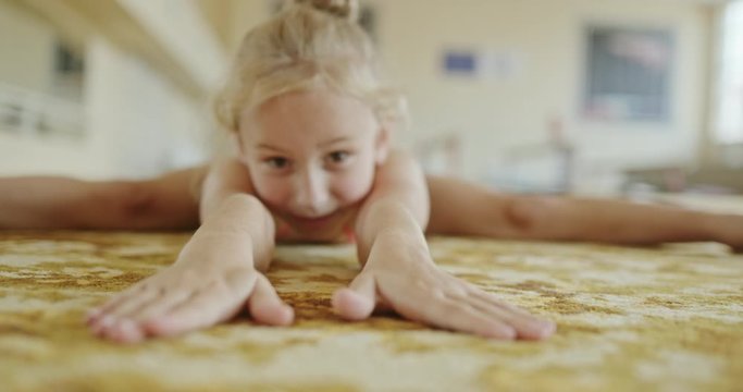 Little girl in leotard bending forward and stretching body while doing split on floor in gym