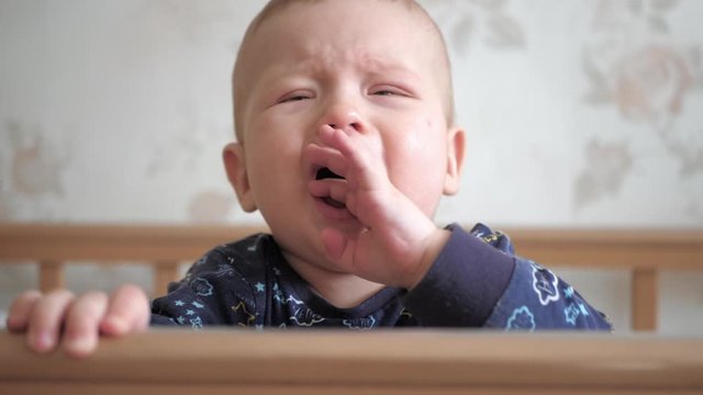 Сute baby with blue eyes stands and screams, freaks out, cries in wooden crib. The child is dressed in blue shirt and holds on to the crib. Slow motion view