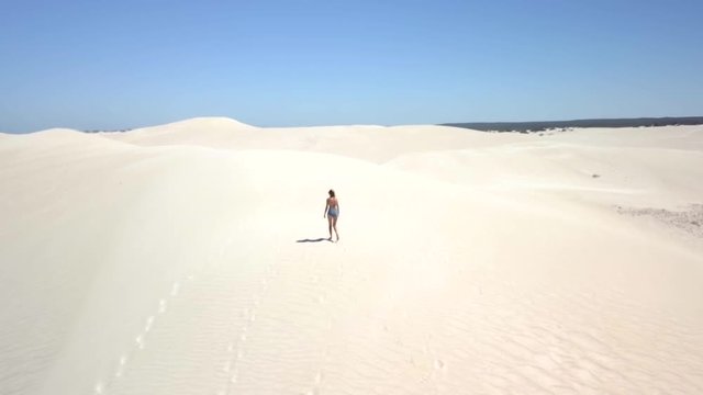 An aerial clip following a girl walking in the sand revealing the sand dunes during mid-day.