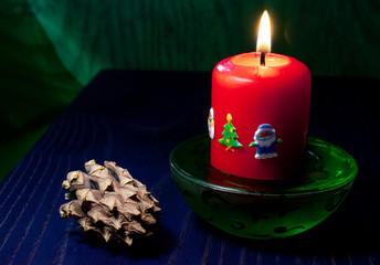Red burning candle with Christmas stickers in a green candlestick on a blue table and a green background next to a cone - Christmas comfort