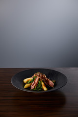 delicious warm salad with arugula, potato and meat in black plate on wooden table on grey background
