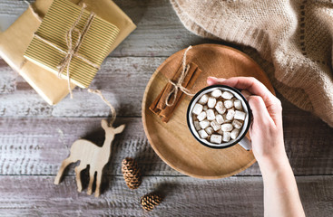 Christmas cocoa in female hand with marshmallow on wooden table background. Top view, flat lay. Holiday drink with festive decoration