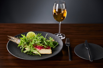 tasty restaurant fish steak with lime and arugula on wooden table near cutlery and white wine on black background