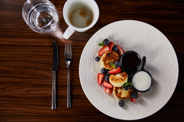 top view of traditional Ukrainian dish syrniki with strawberry, blueberry served with jam and sour cream on wooden table near cutlery, coffee and water