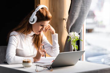 Beautiful thoughtful woman in headphones watching webinar on laptop in cafe