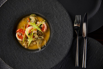 top view of delicious restaurant soup with shrimps in black plate with cutlery