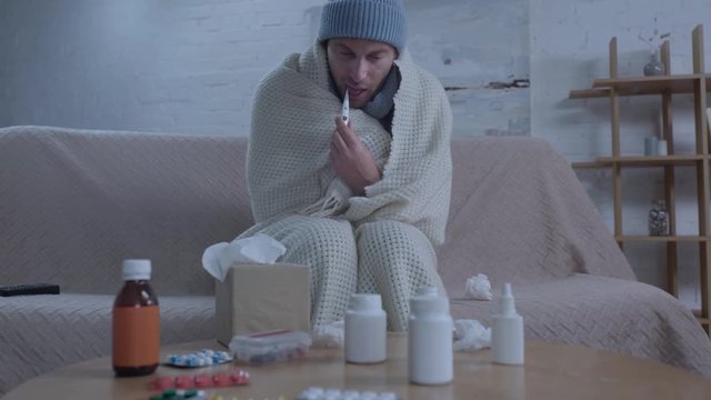 deseased man sitting near table with medicines and measuring temperature