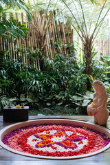 Outdoor jungle view stone round bath tub with flower shaped petals in pink,red,white colors near window. Organic spa relaxation in luxury Bali bathroom.