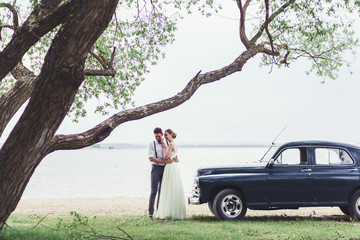 Beautiful wedding couple bride and groom at wedding day outdoors at river beach near old car