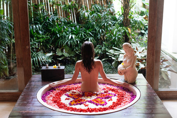 Back view of brunette Young woman in bath with petals of tropical flowers and aroma oils. Spa treatments for skin rejuvenation. Girl relaxing in luxury round bathtub