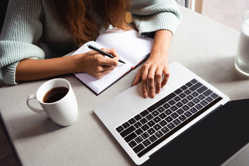 cropped view of woman writing in notepad and using laptop in cafe with cup of coffee