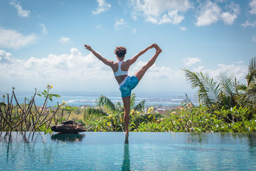 Young woman practicing yoga pose ashtanga hatha asana on the edge of infinity pool with breathtaking view at private villa in Bali