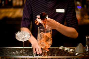Professional bartender pouring a alcoholic drink from the jigger to a cocktail glass with ice cubes