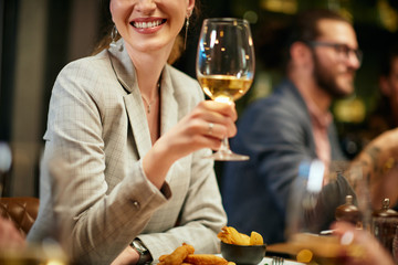 Cropped picture of beautiful caucasian brunette dressed elegant posing with glass of wine while sitting in restaurant with her friends.