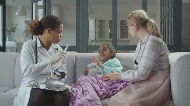Friendly african female pediatrician holding syringe with injection vaccination, consoling together with caring mom and convincing unwell mixed race girl scared of injection to inject medicine at home