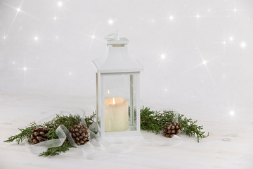 Christmas copy space with a lit candle inside a white lantern, pine branches, white organza ribbon and natural pine cones on a light wooden background with stars light effect