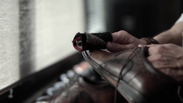 a white man hands in a brown vest white shirt with a bracelet on his hands roll up a rag and clean brown shoes in a dark room