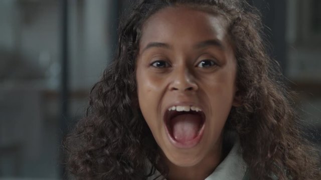 Portrait of lovely elementary age african american girl in casual clothing with long curly hair screaming loud with mouth wide open indoors, expressing negativity, stress and dissatisfaction.