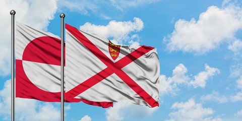 Greenland and Jersey flag waving in the wind against white cloudy blue sky together. Diplomacy...