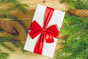 Christmas gift box with a red bow on a wooden background surrounded by fir branches. Close-up, top view. Background about christmas and new year. Frame with fir branches on a wooden background 