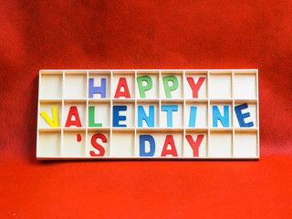 Fototapeta premium Multicolored wooden alphabets or letters of happy valentine 's day texts or words in wooden grids box on rough vivid or vibrant gradient red paper texture and background.