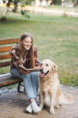 full length view of beautiful young girl in casual clothes reading book and petting golden retriever while sitting on wooden bench in park