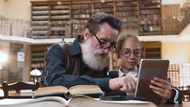 Serious bearded senior grandpa with lovely granddaughter looking at i pad on the library interier background