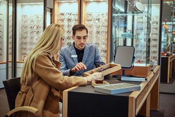 Woman is choosing eyeglasses while male optician sitting near with another eyeglasses.