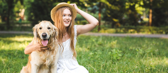 panoramic shot of beautiful young girl in white dress touching straw hat while hugging golden...