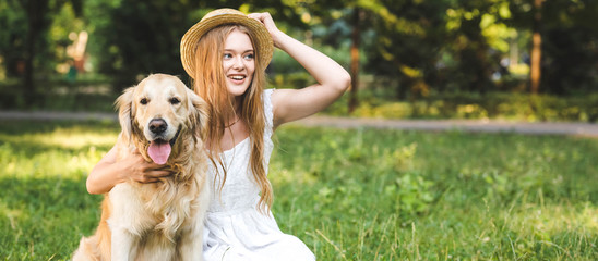 panoramic shot of beautiful young girl in white dress touching straw hat while hugging golden retriever while smiling, sitting on meadow and looking away