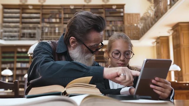 Good-looking eldery bearded grandpa sitting at the table in library with his pretty granddaughter and comparing print book with ebook
