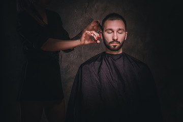 Dark portrait of young gentleman at hairdressing studio, he gets hair trimming from woman.