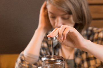 A blonde in a plaid shirt at a table in a cafe brews green tea in a glass teapot.