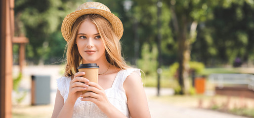 panoramic shot of beautiful girl in white dress and straw hat holding paper cup and looking away
