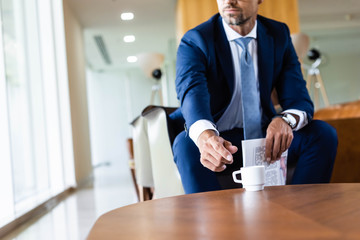 cropped view of businessman in suit taking cup and holding newspaper