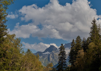 Panoramic view of Mount Tsakhvoa, autumn forest on steep slopes, curved river valley foreground; crones of pine fir trees in warm sunset light; dramatic clouds on snowy peak; natural travel background