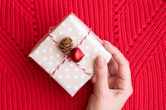 Famale hand holding  a gift decorated with Christmas red ball and natural cone on red Knitting background.Merry Christmas and Napy New Year concept.