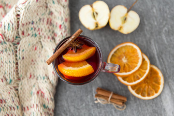 christmas and seasonal treats concept - glass of hot mulled wine with dry orange slicess and cinnamon, star anise, knitted scarf and apples on grey stone surface