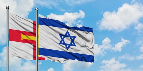 Guernsey and Israel flag waving in the wind against white cloudy blue sky together. Diplomacy concept, international relations.