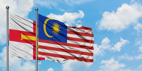 Guernsey and Malaysia flag waving in the wind against white cloudy blue sky together. Diplomacy...