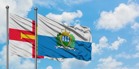 Guernsey and San Marino flag waving in the wind against white cloudy blue sky together. Diplomacy concept, international relations.