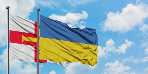Guernsey and Ukraine flag waving in the wind against white cloudy blue sky together. Diplomacy...