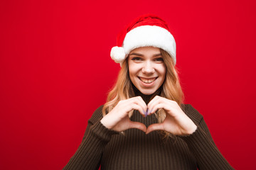 Happy girl in christmas hat shows heart gesture on red background, looks into camera and smiles. Lady in Santa hat shows gesture of her love. Xmas