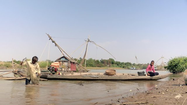 Clammer is shaking a clam net in the water for removing sand from a net and pouring his harvest into a bamboo basket held by his wife sitting on a boat. 