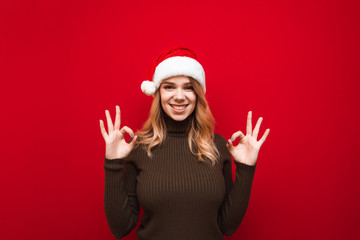 Happy girl in santa hat and warm sweater isolated on red background, hands showing OK gesture,looking into camera and smiling.Studio portrait of joyful lady in christmas hat showing gesture all right