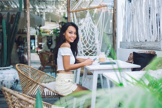 Youthful indonesian woman on terrace outside cafe messaging smiling