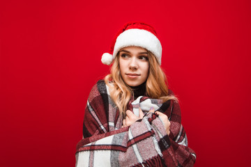 Beautiful blonde girl warms up in plaid on red background, wears santa hat and looks away. Portrait of a frozen girl warming up in plaid, isolated.